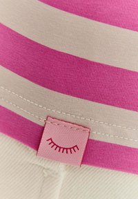 Pruhovaná tkanina s širokými růžovými a béžovými pruhy, s růžovou etiketou, která na lemu zobrazuje jednoduchou grafiku řas. Měkká textura.