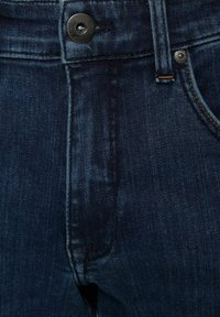 Sötétkék farmernadrág sima felülettel, fém gombbal és kontrasztos varrásokkal a deréknál és a zsebeknél.