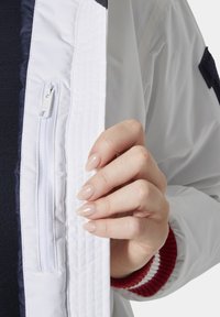 Bílá bomber bunda s kapsou na zip, s námořnickou modrou podšívkou a červenými ribbed manžetami, držena otevřená, aby ukázala vnitřní prvky.