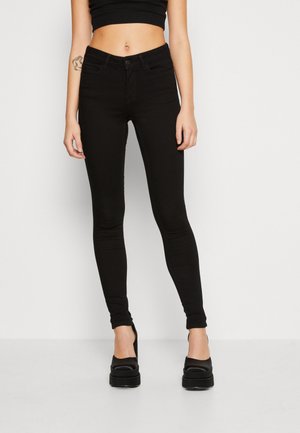 Vaqueros skinny negros de denim, con bolsillos delanteros y traseros, pasadores para cinturón y una silueta ajustada. Combinados con gruesos zapatos de plataforma negros.