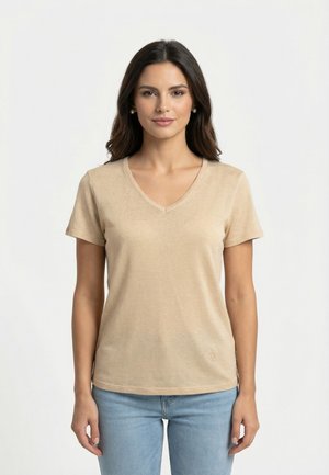 Mujer con cabello largo y oscuro, lleva pendientes de perlas, una camiseta beige de manga corta con cuello en V y vaqueros azul claro, posando contra un fondo blanco.