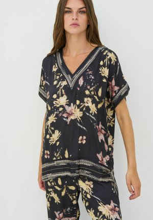 Kvinde iført en sort blomster V-hals top og matchende løse bukser med beige og lyserøde blomster mønstre, der står mod en ensfarvet baggrund.