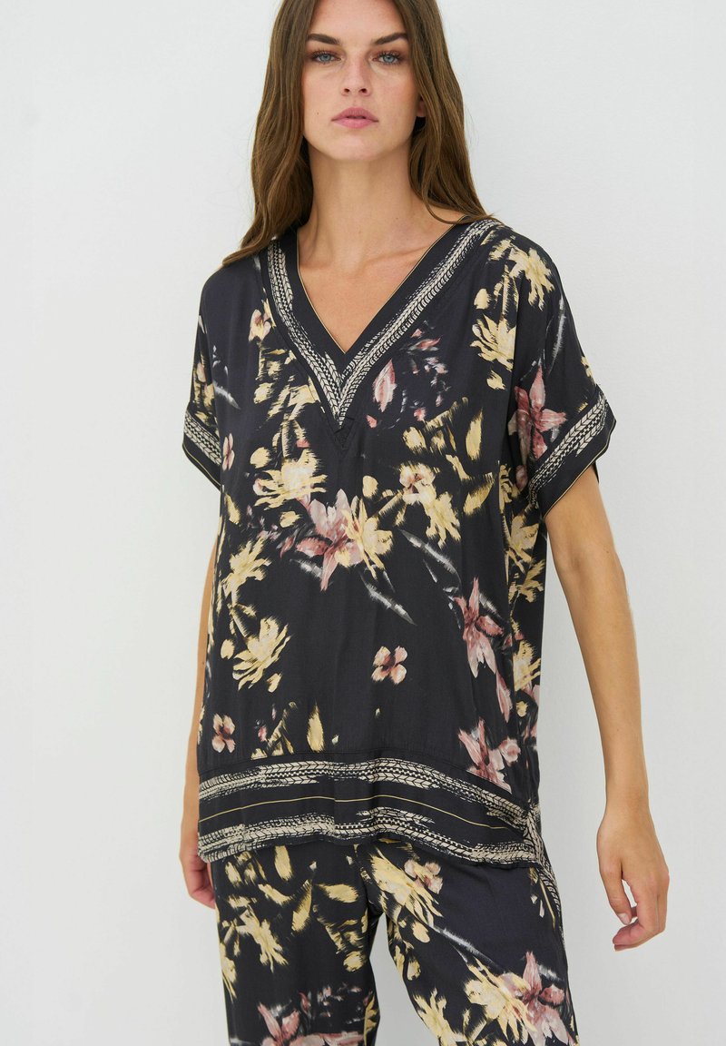 Femme portant un haut noir à fleurs à col en V et un pantalon ample assorti avec des motifs floraux beige et rose, debout devant un fond uni.