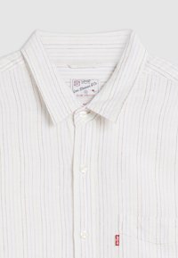 Chemise à rayures blanches avec des lignes rouges et bleues. Comprend un col, une seule poche et une petite étiquette de marque rouge sur la poche.
