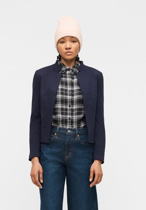 Kvinde iført en lyserød hue, marineblå blazer, sort-hvid ternet skjorte og højtaljede blå jeans, stående mod en hvid baggrund.