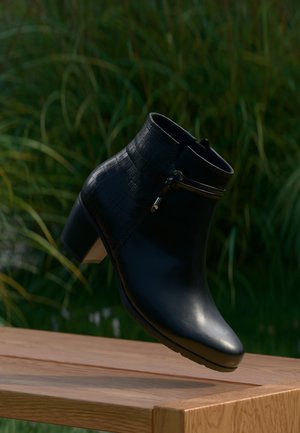 Bottine noire à cheville avec talon bloc bas et partie supérieure texturée, présentée sur un fond vert flou sur une surface en bois.