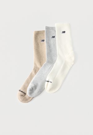 Drie geribbelde crew sokken in beige, grijs en wit, elk met een zwart logo vlakbij de bovenkant en tenen, naast elkaar geplaatst op een effen achtergrond.