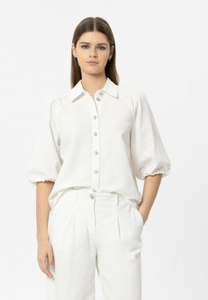 Young woman with straight brown hair wearing a white button-up shirt with puff sleeves and white high-waisted pants, hands in pockets.