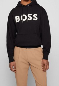 Persona che indossa una felpa nera con la scritta "BOSS" in lettere bianche e pantaloni beige a pieghe, in piedi su uno sfondo neutro.