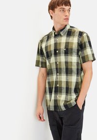 Korte mouw geruite blouse in groen, zwart en lichtbeige met een knoopsluiting kraag en subtiel logo op de borst. Zacht katoenen stof.