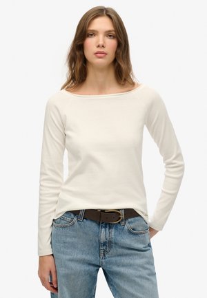 Top crema a maniche lunghe con scollo ampio, abbinato a jeans denim blu chiaro e una cintura marrone con fibbia dorata.