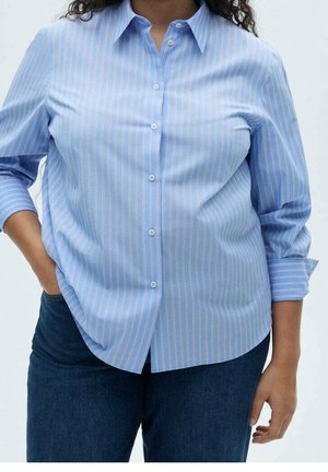 Femme portant une chemise bleue à boutons avec des rayures blanches et jaunes verticales et un jean bleu foncé, main dans la poche, debout devant un fond uni.