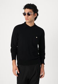 Maglione nero con colletto a polo, maniche lunghe e taschino sul petto. Realizzato in materiale morbido, presenta un dettaglio con bottone dorato.