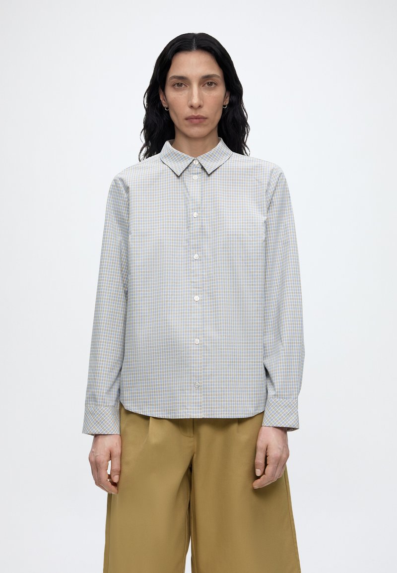 Γυναίκα με μακριά σκούρα μαλλιά φορώντας ένα ανοιχτομπλέ και μπεζ καρό πουκάμισο με κουμπιά και μουσταρδί φαρδιά παντελόνια, που στέκεται μπροστά σε έναν απλό λευκό φόντο.