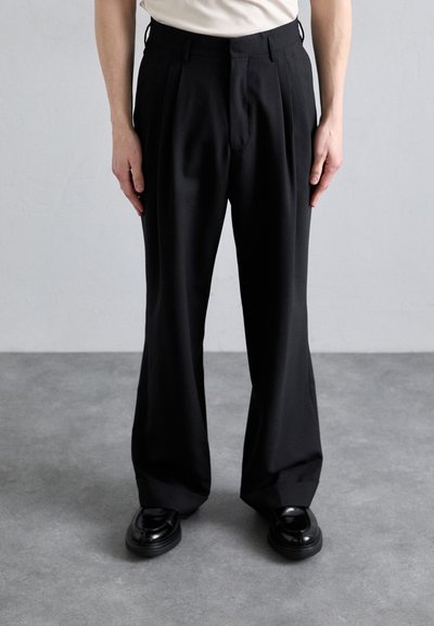 Black tailored trousers with a pleated front. Made from smooth fabric, they feature a classic fit and sit above sleek black shoes.