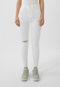 Valkoiset korkeavyötäröiset skinny-farkut, joissa on kuluneet polviyksityiskohdat ja repsottaa helmia, yhdistettyinä vaaleanvihreisiin tennareihin. Sujuva farkkukangas.
