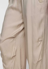 Beige lichte broek met een relaxte pasvorm, voorzien van verticale naden en zijzakken. De stof lijkt glad en licht glanzend.