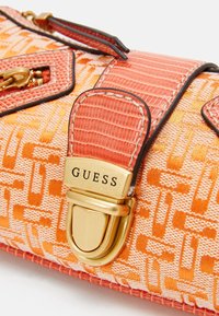 Sac à main en tissu à motif orange avec fermoir doré gravé "GUESS" et détail de bandoulière en cuir orange texturé.