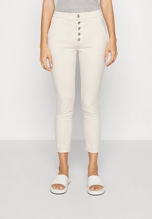 Kobieta ubrana w dopasowane, kremowobiałe spodnie z pięcioma przednimi guzikami oraz białe wsuwane sandały, stojąca na tle jasnego, jednolitego tła.