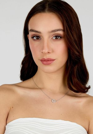 Collana d'argento con un pendente circolare che presenta piccoli gemme trasparenti, indossata sulla pelle nuda sotto un morbido tessuto bianco testurizzato.