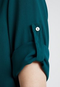 Turkusowa bluzka z podwiniętymi rękawami, z detalem guzikowym. Wykonana z gładkiego materiału o miękkiej teksturze, charakteryzująca się prostym wzornictwem.