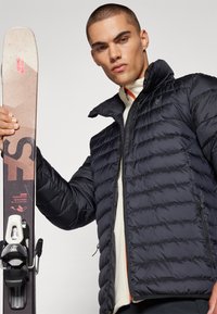Dämpfjacke in Marineblau mit gestepptem Muster, hohem Kragen und Reißverschlusstaschen. Das Modell hält einen Ski mit einem beige-schwarzen Design.