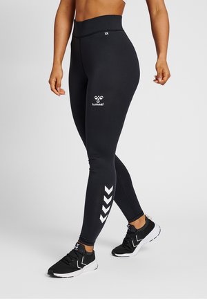 Vrouw draagt zwarte legging met hoge taille, witte Hummel-logo en chevronstrepen, gecombineerd met zwart-witte sneakers, staand met één voet naar voren.