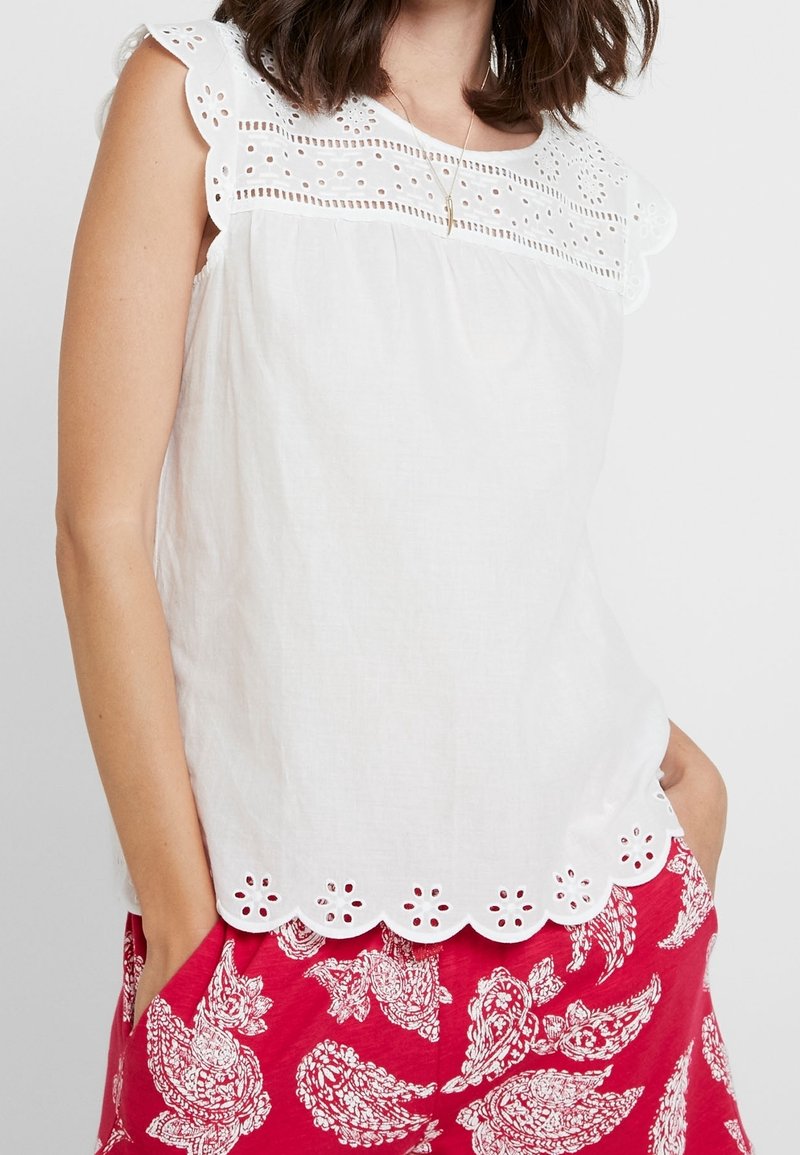 Femme portant un chemisier blanc sans manches avec des détails en broderie anglaise et des bords festonnés, associé à un pantalon rouge à motif cachemire.