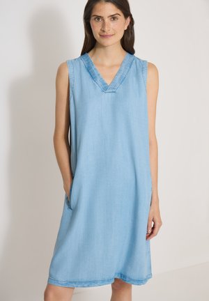 Femme portant une robe en jean bleu clair sans manches avec un col en V et les mains dans les poches, se tenant devant un fond clair uni.