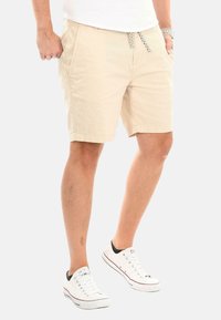 Beige Baumwollshorts mit elastischem Bund, Zugband, seitlichen Taschen und lockerer Passform, kombiniert mit weißen Sneakers.