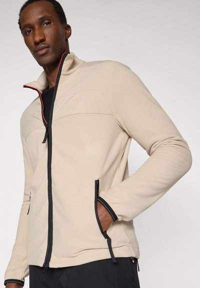 Mann trägt beige Fleecejacke mit Reißverschluss und schwarzem Besatz, Hände in den Taschen, steht vor einfachem hellem Hintergrund.