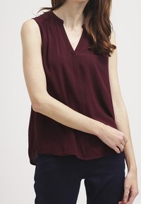 Burgundy sleeveless blouse with a V-neckline, pleated shoulder detailing, and a relaxed fit, made of soft, flowing fabric.