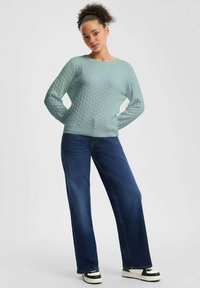 Lichtblauwe gebreide trui met een gestructureerd honingraatpatroon, ronde halslijn en lange mouwen, gecombineerd met wijde donkere blue jeans en sneakers.