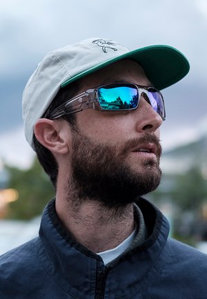 Homme barbu portant des lunettes de soleil bleues réfléchissantes et une casquette blanche à visière verte, regardant vers le haut à l'extérieur sous une lumière douce.