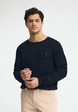 Mann, der den Ärmel eines dunkelblauen Pullover mit Zopfmuster anpasst, trägt beige Hose und steht vor weißem Hintergrund.