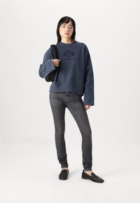 Sweatshirt marine avec un emblème texturé, coupe décontractée et manches larges, associé à un jean sombre et des mocassins noirs. Sac tissé noir en accent.