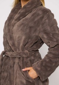 Bata de felpa marrón con una textura suave, cuello de chal, cintura con cinturón y bolsillos laterales. La tela parece cálida y acogedora.