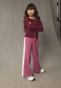 Vinrød ribstrikket cardigan med knapper og lyserøde vidbenede sweatpants med sidestriber, kombineret med hvide træningssko mod en grå baggrund.