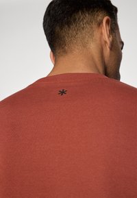 T-shirt rossa di cotone con scollatura rotonda, dotata di un piccolo logo a stella nera sul retro vicino al colletto. Tessuto liscio, design semplice.