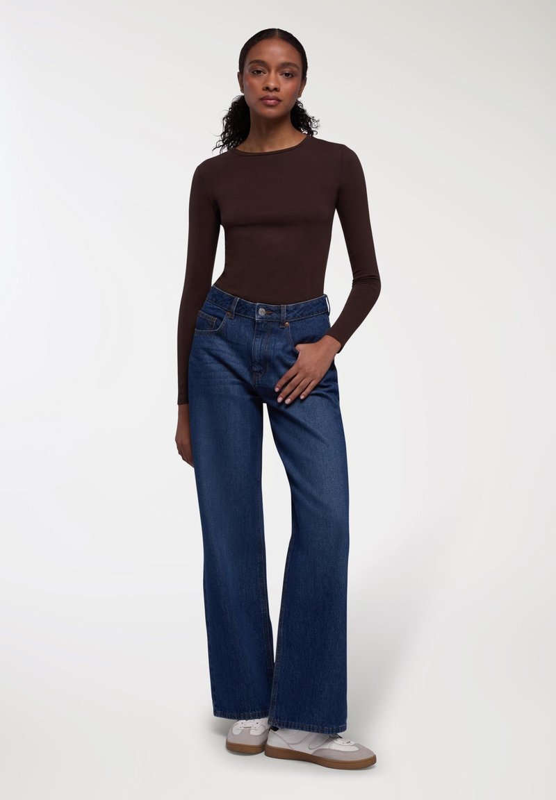 Top marrone a maniche lunghe abbinato a jeans blu in denim a vita alta e gamba larga. Il modello indossa sneaker bianche e grigie. Sfondo semplice.