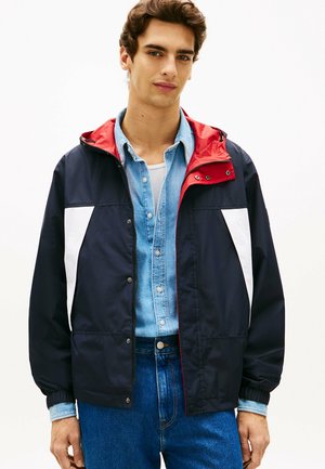 Ung mand iført en marineblå jakke med hvide paneler og rød hætteforing over en denimskjorte og hvid undertrøje, stående mod en ensfarvet baggrund.