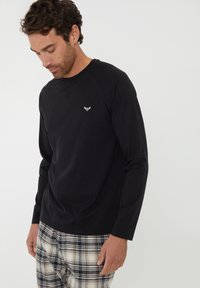 T-shirt noir à manches longues avec un col rond et un logo discret. Assorti avec un pantalon de pyjama à carreaux dans des tons blanc, noir et beige.