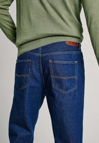 Jeans de mezclilla azul con corte recto, con costuras naranjas, dos bolsillos traseros y una etiqueta de cuero marrón en la cinturilla.
