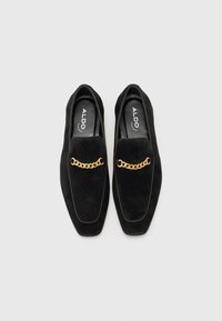 Paire de mocassins en daim noir avec détail de chaîne dorée sur le devant, semelles intérieures marquées ALDO, posés sur un fond blanc uni.