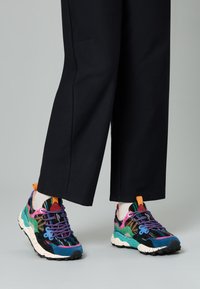 Des baskets colorées présentent un mélange de textures en violet, vert, rose et bleu, avec une semelle mouchetée et des lacets lumineux.