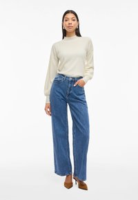 Cream, high-neck sweater with long sleeves paired with high-waisted, wide-leg blue jeans and brown pointed heels, set against a white background.