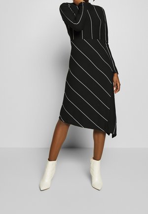 Woman wearing a black long-sleeve dress with white vertical and diagonal stripes, paired with white ankle boots, standing on a gray floor.