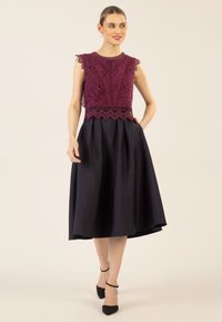 Ärmelloses Kleid mit einem bordeauxroten Spitzenoberteil mit gewellten Kanten und einem marineblauen Rock. Der Rock hat Taschen und eine ausgestellte Silhouette.