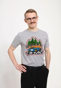 Camiseta gris de manga corta con un diseño gráfico de árboles verdes y un coche azul. El texto en negro y rojo resalta el diseño.