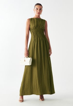 Olivegrünes Maxikleid mit strukturierter Oberfläche, das vertikale Falten und einen Bootsausschnitt aufweist. Kombiniert mit einer weißen, gesteppte Handtasche.